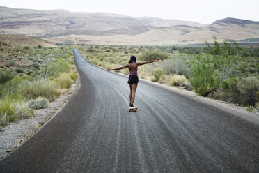 Rear view of woman skateboarding on road amidst field - CAVF22271