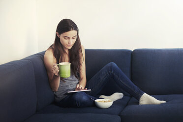 Frau, die ein digitales Tablet benutzt, während sie auf dem Sofa zu Hause frühstückt - CAVF22333