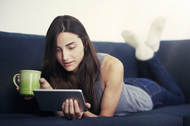 Junge Frau hält eine Kaffeetasse und benutzt ein digitales Tablet auf dem Sofa - CAVF22336