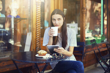 Frau trinkt Kaffee, während sie ein digitales Tablet hält und in einem Straßencafé sitzt - CAVF22339