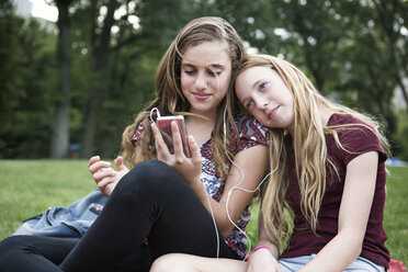 Smiling sisters listening music while relaxing on grassy field at park - CAVF22379
