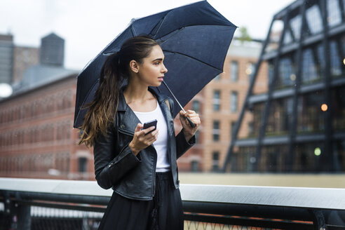 Woman carrying umbrella and using phone while standing at railing in city - CAVF22392