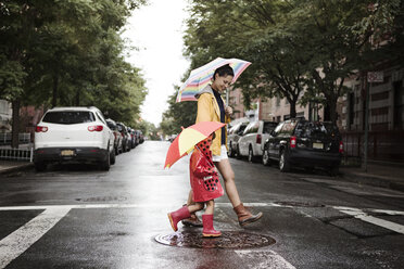 Seitenansicht von Mutter und Tochter mit Regenschirmen beim Überqueren der Straße - CAVF22409