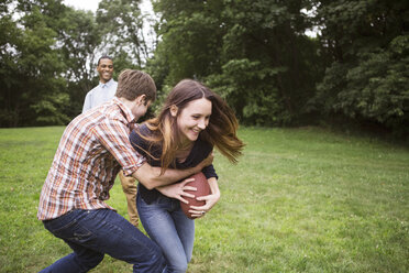 Mann zieht Frau zurück, die einen Fußball hält, während ein Freund auf einer Wiese steht - CAVF22428