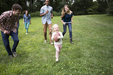 Verspielte Freunde laufen hinter einem Mädchen, das einen Fußball auf einer Wiese hält - CAVF22429