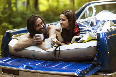 Cheerful couple holding coffee mugs while lying in pick-up truck at forest - CAVF22468