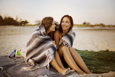 Lächelnde Frauen in Decke eingewickelt am Strand sitzend - CAVF22491