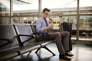 Geschäftsmann, der sein Smartphone benutzt, während er auf einem Sitz im Warteraum eines Bahnhofs sitzt - CAVF22515