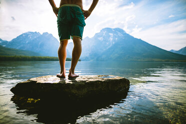 Low section of man with hands on hip standing on rock in lake against mountains - CAVF22617