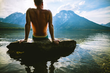 Mann ohne Hemd entspannt sich auf einem Felsen im See vor Bergen - CAVF22618