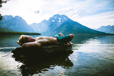 Mann ohne Hemd, der auf einem Felsen im See liegt, während er sich vor den Bergen entspannt - CAVF22620