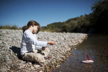 Girl pulling toy boat with rope while sitting at lakeshore on sunny day - CAVF22718