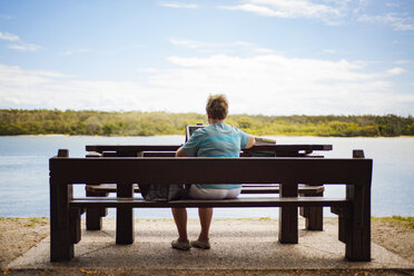 Rückansicht einer älteren Frau, die einen Laptop benutzt, während sie auf einer Bank am Seeufer gegen den Himmel sitzt - CAVF22763