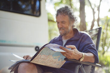 Glücklicher älterer Mann, der auf einem Stuhl im Wald sitzend ein Kartenbuch liest - CAVF22769