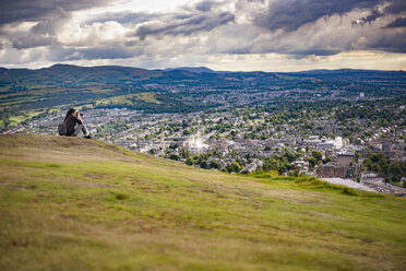 Young woman photographing while sitting on hill by cityscape against cloudy sky - CAVF22780