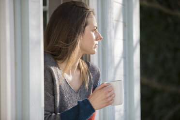 Nachdenkliche Frau hält Tasse am Fenster stehend - CAVF22918