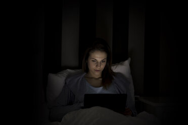 Woman using laptop computer while lying on bed in darkroom - CAVF22920