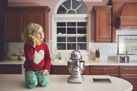 Nettes Mädchen spielt mit Banane am Tisch bei Gerät in Küche, lizenzfreies Stockfoto