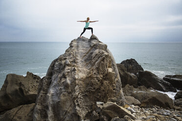 Woman practicing worrier 2 pose on rocks by sea - CAVF22982