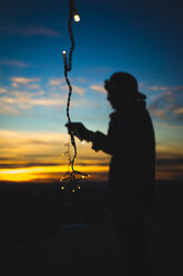 Silhouette man holding fairy lights while standing on field against sky during sunset - CAVF23069