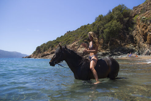 Woman wearing bikini sitting on horse in river against clear sky - CAVF23078