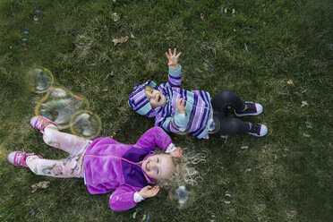 Draufsicht auf Geschwister, die mit Seifenblasen spielen, während sie auf einer Wiese liegen - CAVF23086