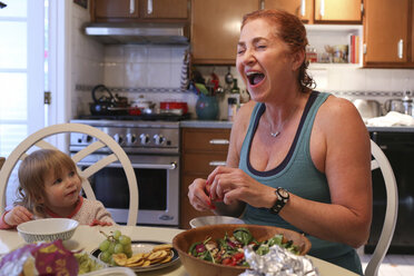 Cheerful grandmother preparing food while sitting with granddaughter at table - CAVF23124