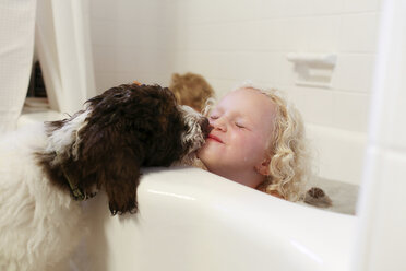 Hund küsst Mädchen, das in der Badewanne im Badezimmer sitzt - CAVF23127