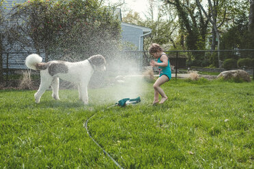 Side view of girl and dog standing by spraying water at yard - CAVF23153