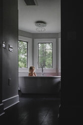 Girl looking away while sitting in bathtub against windows seen through doorway - CAVF23173