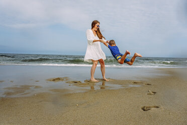 Playful mother swinging son on shore at beach against sky - CAVF23232