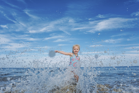 Porträt eines verspielten Mädchens, das im Meer stehend gegen einen bewölkten Himmel Wasser spritzt, lizenzfreies Stockfoto