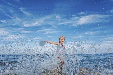 Porträt eines verspielten Mädchens, das im Meer stehend gegen einen bewölkten Himmel Wasser spritzt - CAVF23237