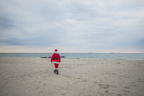 Rückansicht eines älteren Mannes im Weihnachtsmannkostüm, der am Strand spazieren geht - CAVF23254