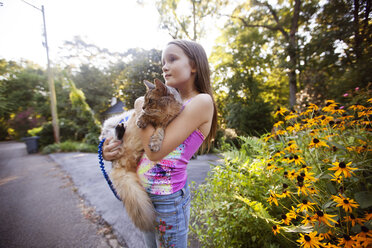 Mädchen trägt Katze, während sie auf der Straße gegen Pflanzen steht - CAVF23302