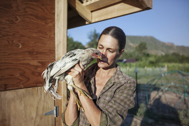 Frau hält ein Huhn, während sie an der Scheune eines Bauernhofs steht - CAVF23311