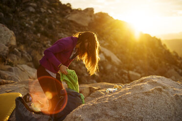 Side view of woman packing bag on rocks against sky during sunset - CAVF23451