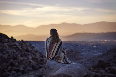 Rückansicht einer in eine Decke eingewickelten Frau, die auf einem Felsen gegen den Himmel sitzt - CAVF23459