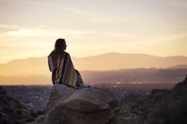 Rear view of woman wrapped in blanket sitting on rock against sky during sunset - CAVF23460