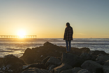 Side view of woman standing on rocky shore against sky during sunset - CAVF23463