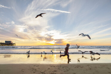 Side view of girl playing with seagulls at beach against sky during sunset - CAVF23515