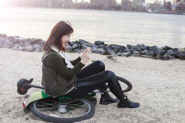 Seitenansicht einer glücklichen Frau, die ihr Smartphone benutzt, während sie auf einem Fahrrad am Strand sitzt - CAVF23535