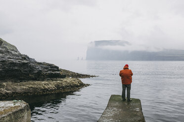 Rear view of hiker in hooded jacket standing on pier against sea during foggy weather - CAVF23554