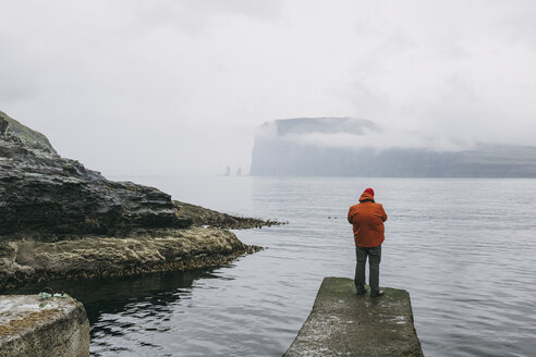 Rückansicht eines Wanderers in einer Kapuzenjacke, der bei nebligem Wetter auf einem Pier am Meer steht - CAVF23554