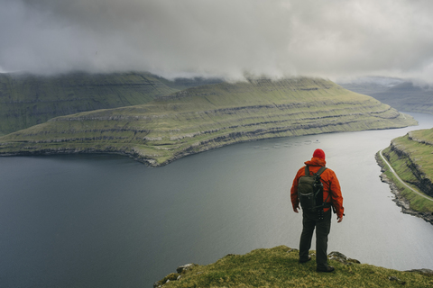 Wanderer mit Rucksack, der die Aussicht betrachtet, während er auf einer Klippe gegen stürmische Wolken steht, lizenzfreies Stockfoto