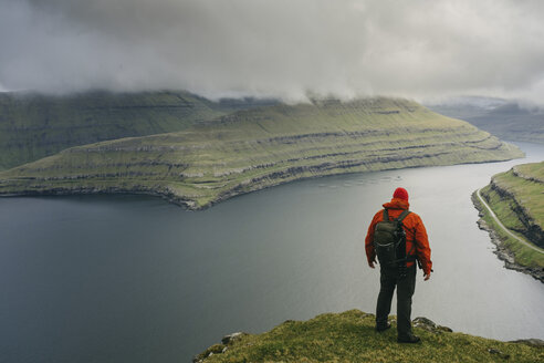 Wanderer mit Rucksack, der die Aussicht betrachtet, während er auf einer Klippe gegen stürmische Wolken steht - CAVF23584