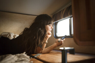 Frau entspannt sich auf dem Bett am Fenster, während sie ihr Smartphone im Wohnmobil benutzt - CAVF23681