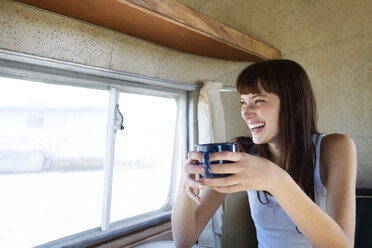Fröhliche Frau trinkt Kaffee, während sie am Fenster eines Wohnmobils sitzt - CAVF23747