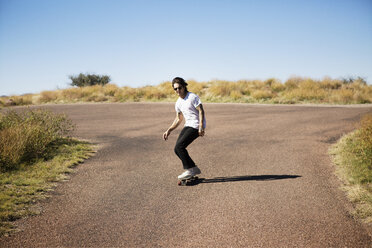 Man skateboarding on road by field against clear sky - CAVF23781