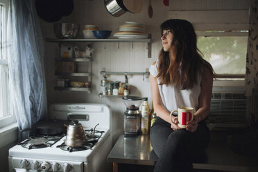 Nachdenkliche Frau hält eine Tasse, während sie auf dem Küchentisch sitzt - CAVF23921